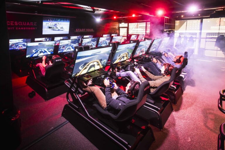 Indoor racing simulator room with multiple high-tech racing seats and screens, featuring Grand Prix Racewear branding, vibrant lighting, and a smoky atmosphere.