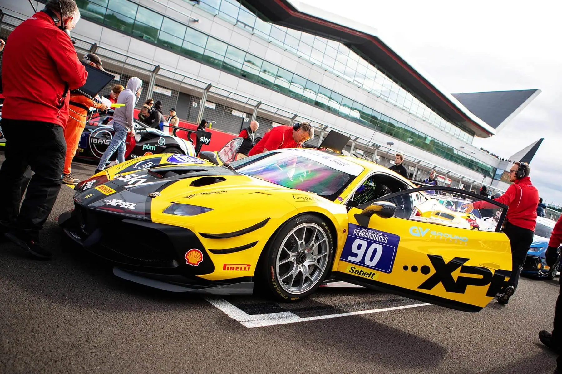 Yellow and black race car with number 90 on the track at a racing event, surrounded by team members and officials.
