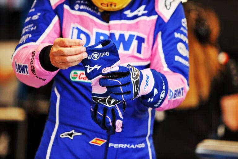 Close-up of a Formula 1 driver wearing blue racing gloves and a racing suit with sponsor logos, preparing for a race.