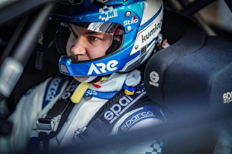 Close-up of a race car driver wearing a blue and white helmet and racing suit, seated in the cockpit, ready for a race.