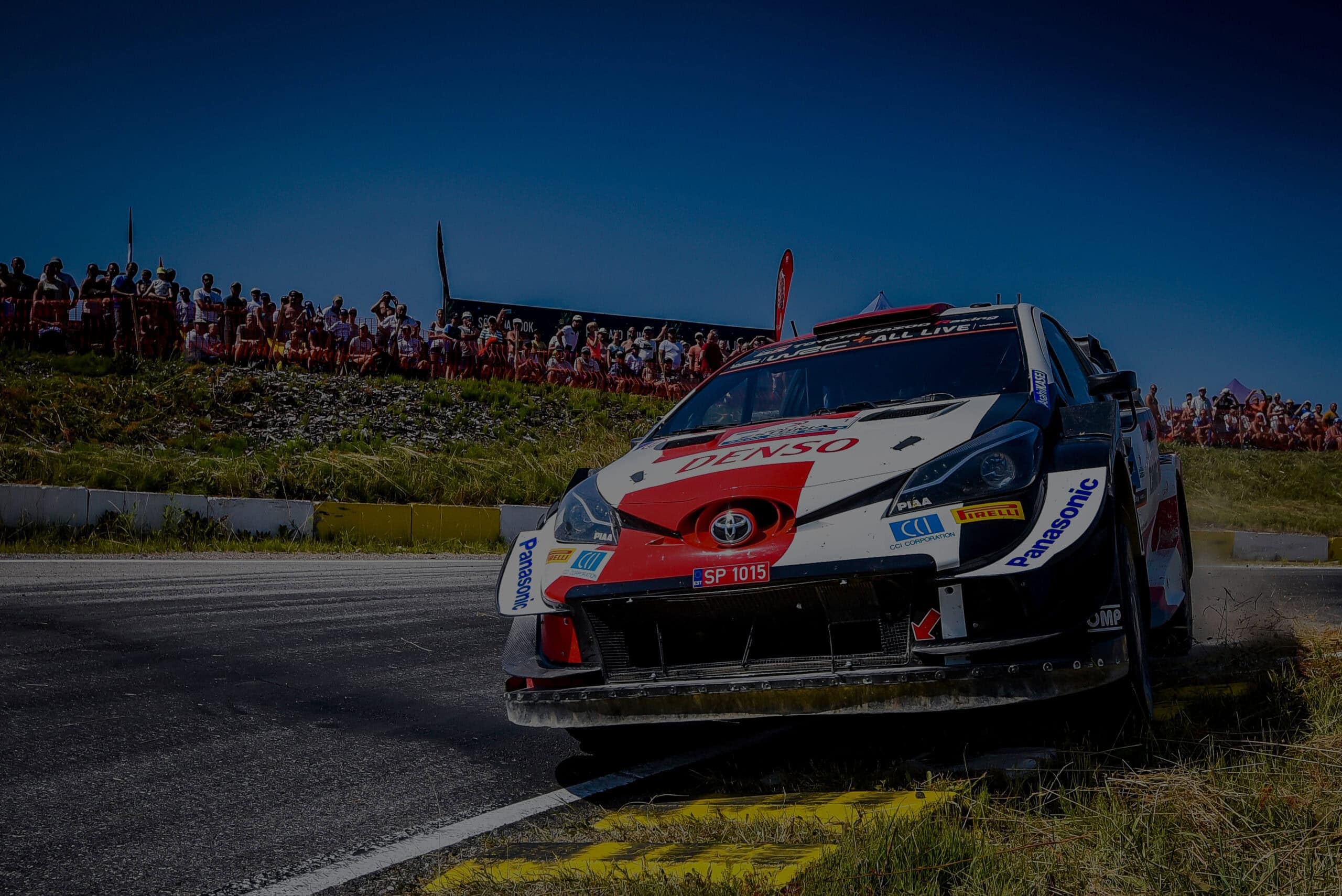 Toyota rally car racing on a dirt track with spectators in the background, showcasing Grand Prix Racewear.