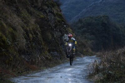 Motorcycle rider navigating a rugged mountain trail during daytime, surrounded by lush greenery and steep rocky slopes.