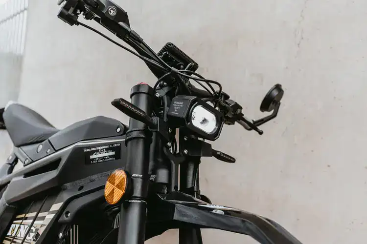 Close-up of a black electric motorcycle with handlebar controls and a digital display, set against a plain wall.