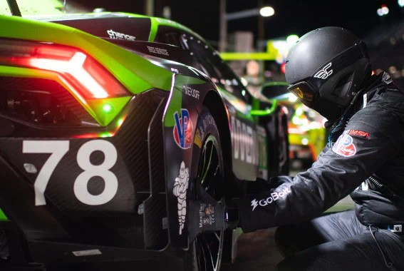Close-up of a green and black Grand Prix Racewear racing car with a mechanic working on the rear wheel at night.