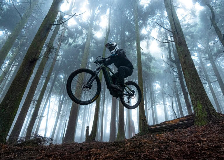Mountain biker wearing protective gear jumps over a forest trail in a misty woodland setting.
