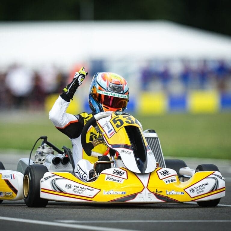 Race car driver in Grand Prix Racewear celebrating victory in a go-kart race, wearing a helmet and racing suit with sponsor logos.