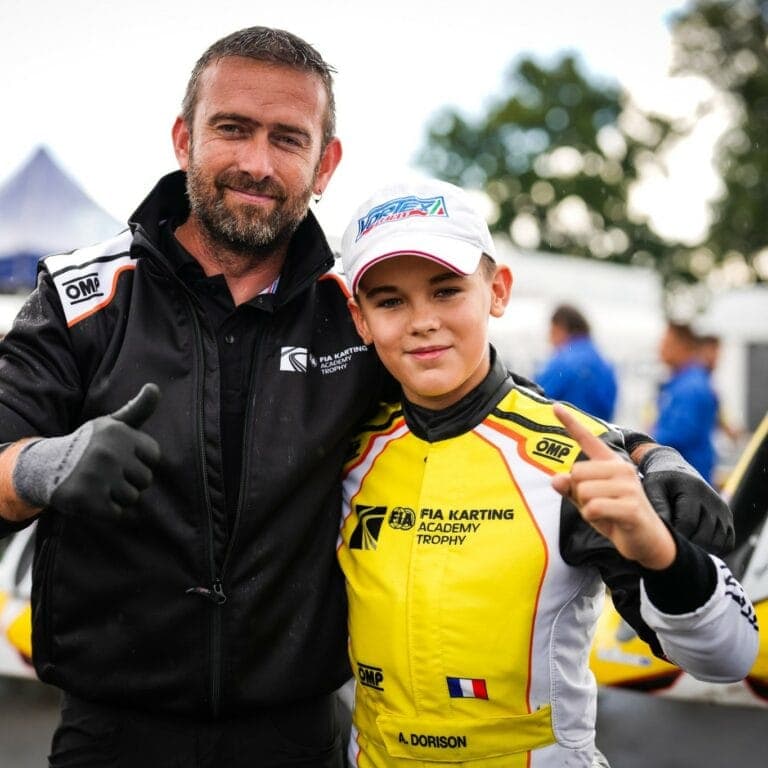 Young karting driver in yellow racing suit with FIA Karting Academy Trophy logo, posing with an adult at a racing event.