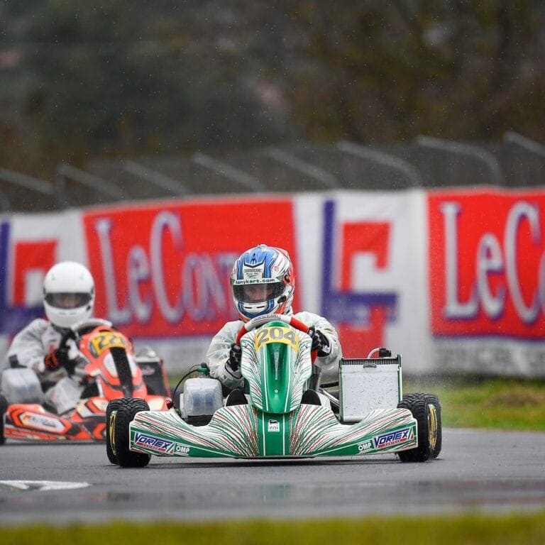 Go-kart racing at a circuit with a driver in a blue helmet and a green and white kart, featuring LeCont branding in the background.