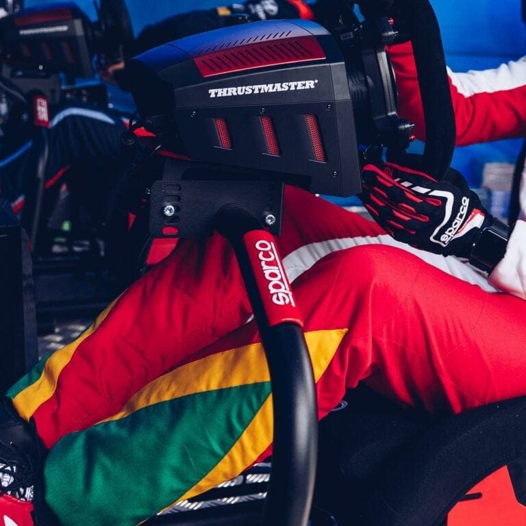 Close-up of a racing driver in a red, yellow, and green suit with a Thrustmaster racing wheel and Sparco gloves, preparing for a race at Grand Prix Racewear.