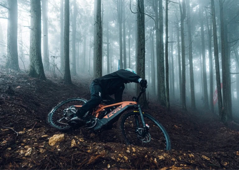 Mountain biker navigating a steep, muddy forest trail in foggy conditions, wearing protective gear and riding a high-performance mountain bike.