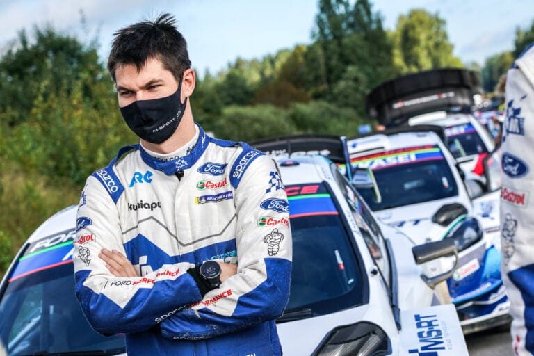 Race car driver in racing suit and face mask standing in front of rally cars during a race event.