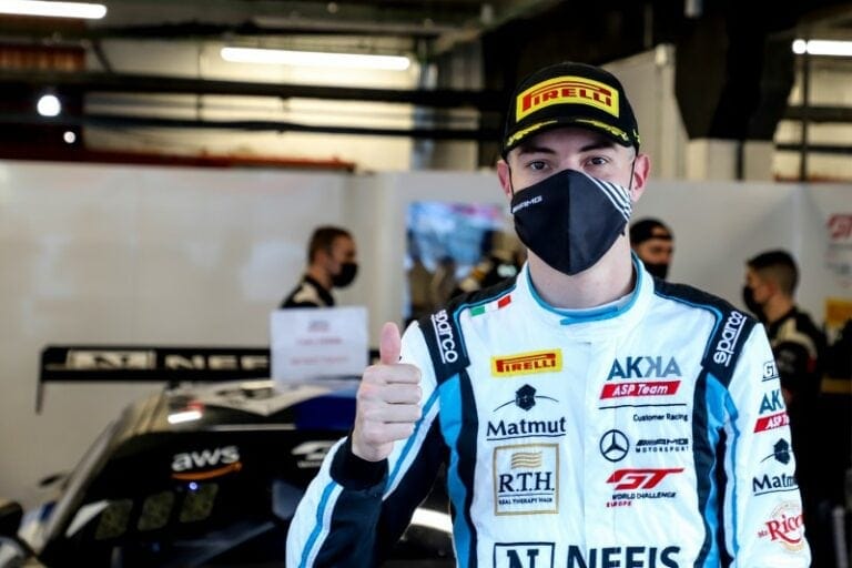 Race car driver in Grand Prix Racewear giving a thumbs up at the race track, wearing a black face mask and racing suit with sponsor logos.