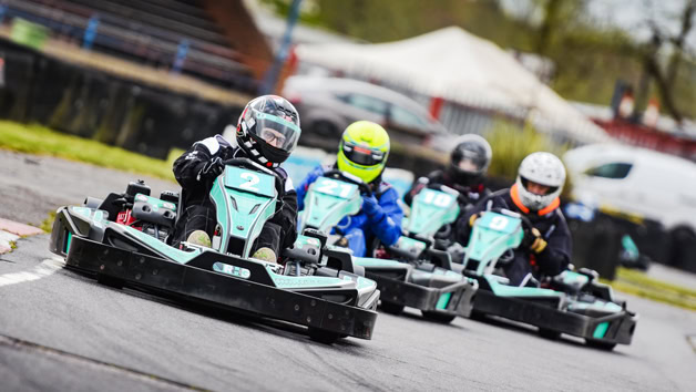 Four go-kart racers wearing helmets and racing suits compete on an outdoor track, demonstrating high-speed karting action.