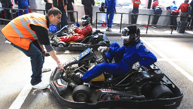 Young man in orange safety vest assisting a driver in a go-kart at a racing event, with other racers and spectators in the background.
