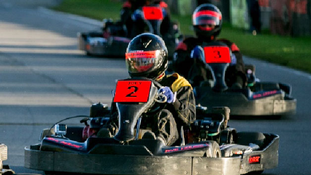 Group of young go-kart racers wearing helmets and racing suits on a track, with the leading driver marked with a red number 2.