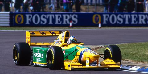Yellow and green vintage Formula 1 race car on track at a motorsport event, with Foster's advertising banners in the background.