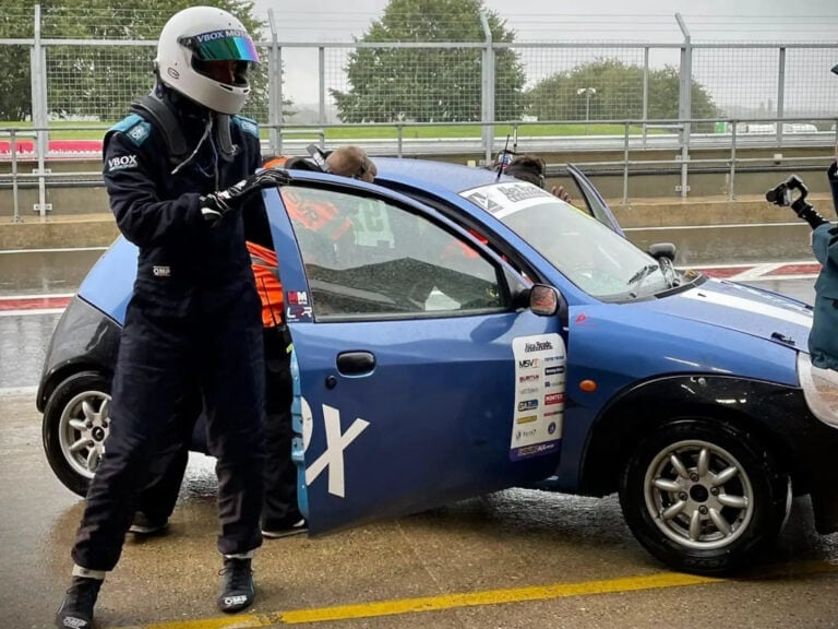Race car driver in full gear inspecting a blue race car at a racing circuit.