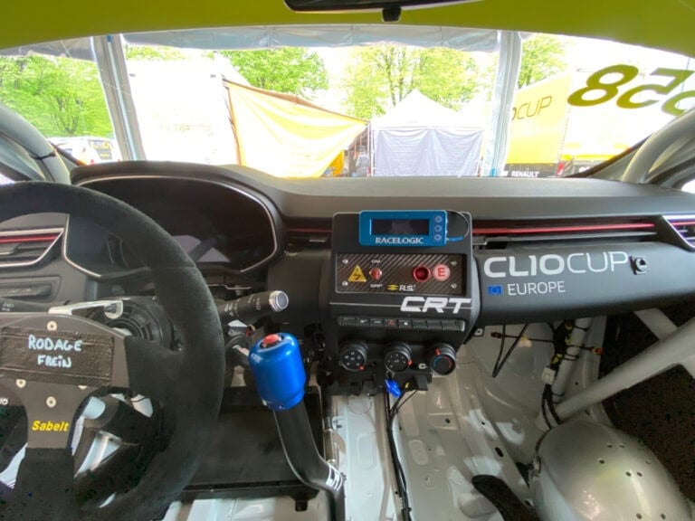 Interior of a race car featuring a digital dashboard, racing steering wheel, and safety equipment, with a focus on Grand Prix Racewear gear and racing technology.