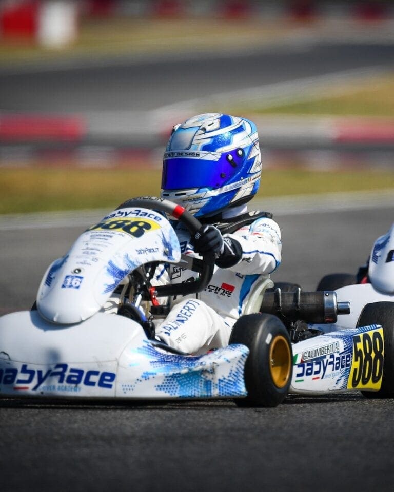 Close-up of a go-kart driver wearing a blue and white helmet and racing suit on a race track.