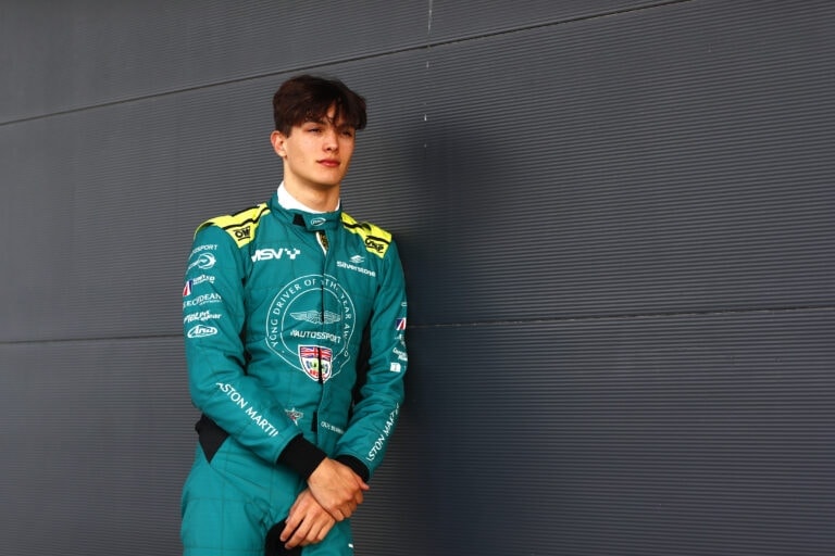 Young race car driver in Grand Prix Racewear standing against a modern grey wall, showcasing professional motorsport apparel.