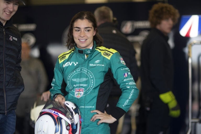 Young female race car driver in racing suit holding helmet at Grand Prix Racewear event.