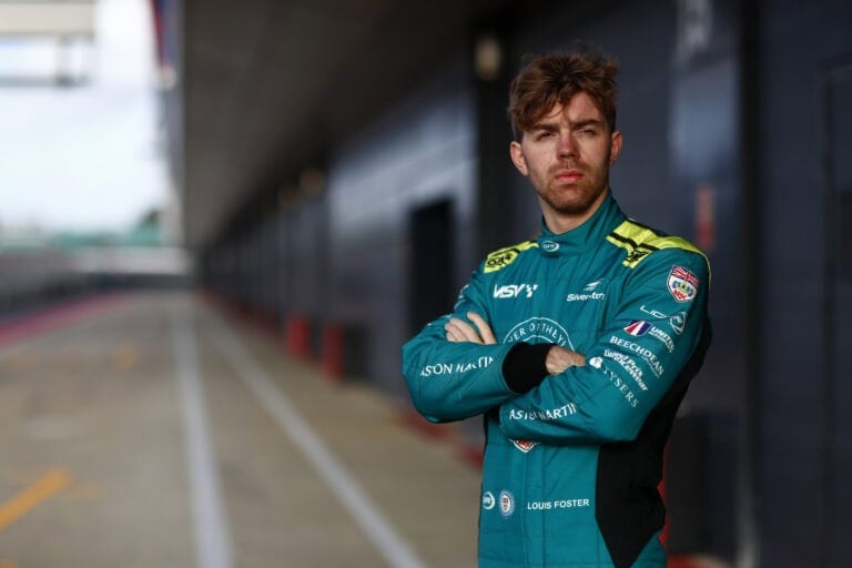 Young male race car driver in blue racing suit standing with arms crossed at a racing circuit.