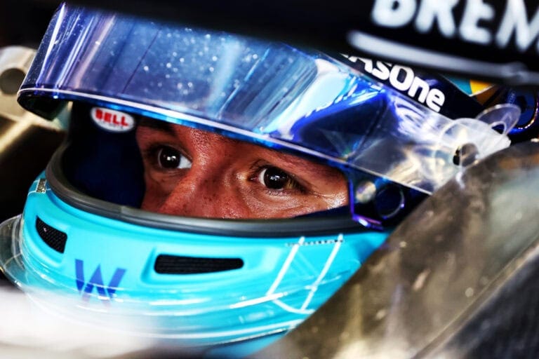 Close-up of a race car driver wearing a blue Bell helmet with a clear visor, focused eyes visible, in a racing environment.