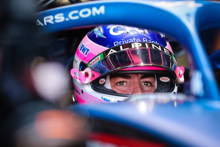 Close-up of a race car driver wearing a Bell helmet with pink accents, sitting in the cockpit of a racing car.