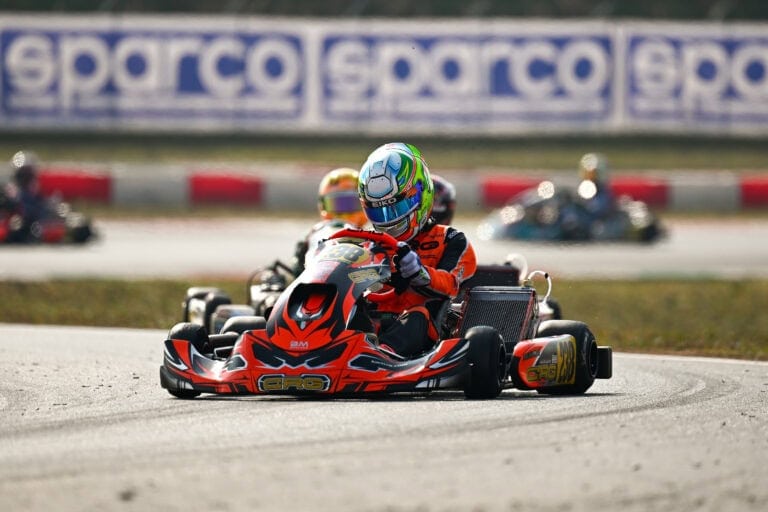 Karter in a red and black racing suit driving a go-kart on a race track during a kart racing event.