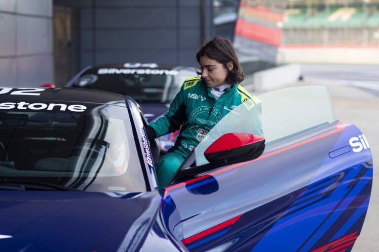 Female race car driver in green racing suit preparing in a blue race car at a track day event.