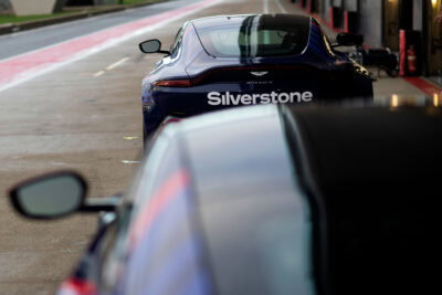 Close-up of a dark blue Silverstone race car in a pit lane, with another vehicle in the foreground, at a racing circuit.