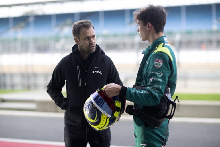 Race car driver in green racing suit with helmet, talking to a team member at a race track.
