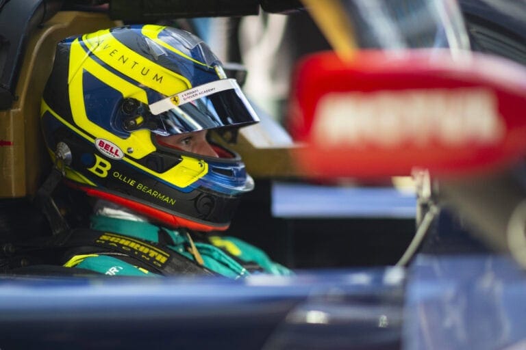 Close-up of a race car driver wearing a yellow and blue helmet with sponsor logos, seated in a race car ready for a track day.