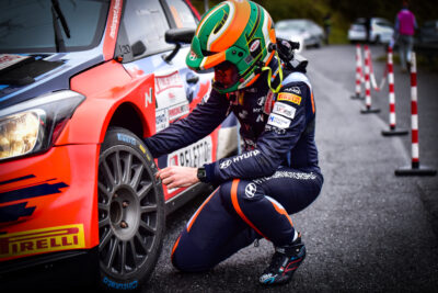 Race car driver in full racing gear inspecting a rally car during a track day event.