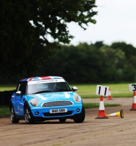Mini Cooper driving on a race track during a motorsport event, with traffic cones and a fallen cone in the foreground.