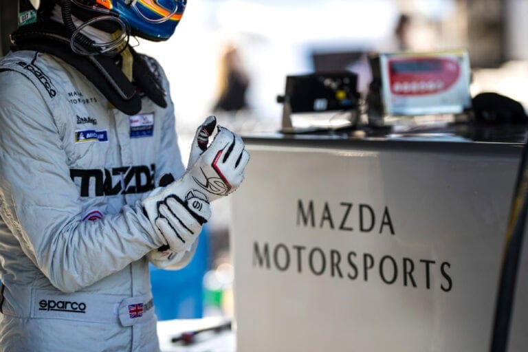 Racing driver in a white suit and gloves preparing for a race at Mazda Motorsports event.