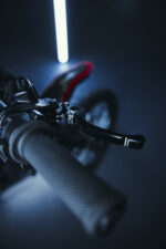 Close-up of a mountain bike handlebar with a textured grip and gear shifter, illuminated by a vertical blue light in a dark setting.
