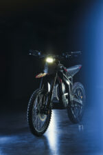 Electric dirt bike with LED headlight on a dark background, showcasing Grand Prix Racewear gear and accessories.