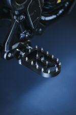 Close-up of a motorcycle footpeg with a textured surface for grip, mounted on a black frame.