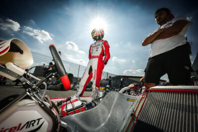 Karting driver in racing suit and helmet standing next to go-kart on race track with sunny sky.