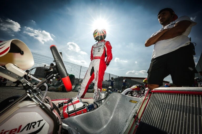 Karting driver in racing suit and helmet standing next to go-kart on race track with sunny sky.
