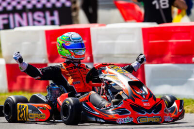 Kart racing driver celebrating victory in a go-kart race, wearing a colourful helmet and racing suit, with a finish line and barriers in the background.