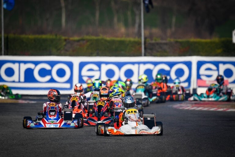 Close-up of go-kart racers competing on a track during a Grand Prix event, showcasing vibrant racing suits and karts with a blurred background.