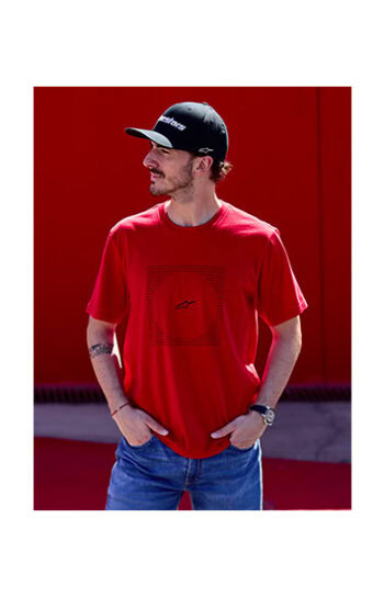 Young man wearing a red Alpinestars racing T-shirt, black cap, and jeans, standing outdoors against a red background.