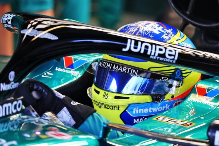 Close-up of a Formula 1 driver in a racing helmet and suit inside a race car, showcasing Grand Prix Racewear gear and branding.