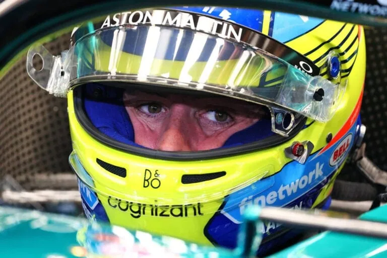 Close-up of a race car driver wearing a yellow and blue helmet with the Astom Martin logo, preparing for a race.