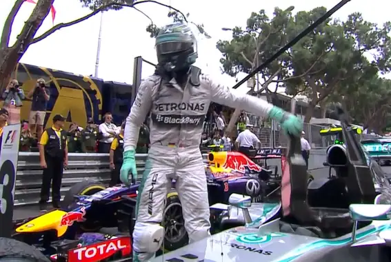 Formula 1 driver in Petronas Mercedes racing suit celebrating on the podium with a trophy, surrounded by team members and race cars.