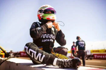 Race car driver in full gear, sitting on the track with helmet and racing suit, preparing for a race at a motorsport event.