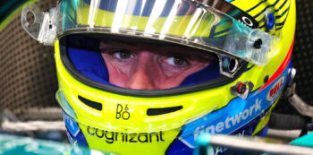 Close-up of a race car driver wearing a bright yellow helmet with blue and black accents, focused eyes visible through the visor.