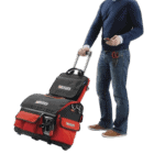 Man with a red and black racing gear bag on wheels, holding a smartphone, wearing casual jeans and a navy sweater.
